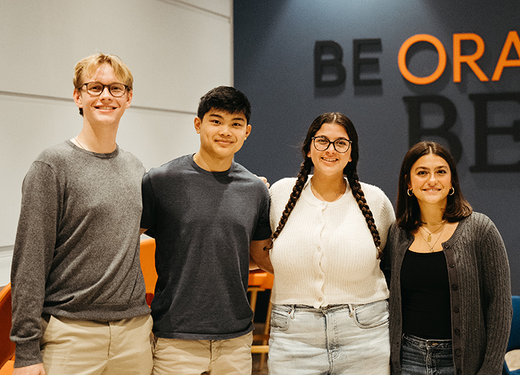 Group of undergraduate students posing in front of wall graphic saying be orange be whitman
