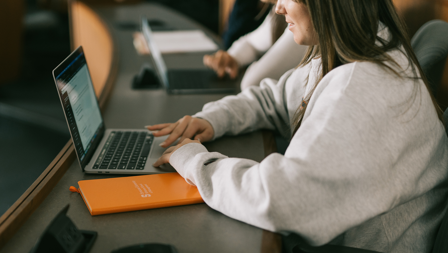 Students working on laptop with orange notebook next to her